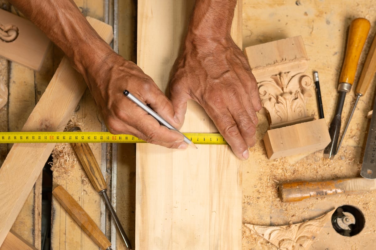 Mains d'un charpentier traçant une coupe au crayon sur une planche, mètre ruban et gouges posés sur l'établi